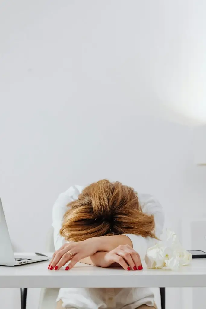 A tired woman with red nails resting her head on a white desk in a bright office setting.